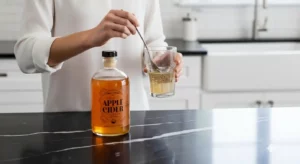 Lemon water or ACV preparation, woman stirring apple cider vinegar in a glass of water on black marble, bottle of ACV in front, white blouse, modern kitchen