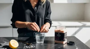 Woman preparing Pink Salt Trick Recipes with Himalayan pink salt, lemon, and water on a black marble countertop in a modern kitchen.